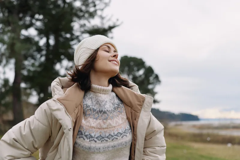Woman outdoors wearing a warm coat and knit sweater, smiling with arms outstretched in a windy countryside landscape under soft skies