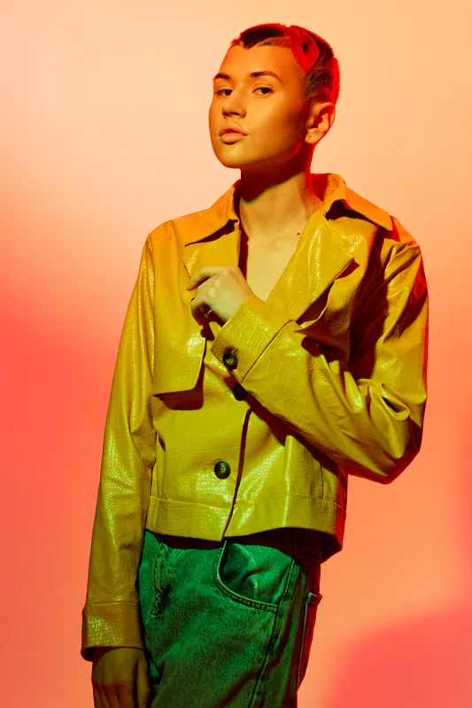 Young woman in yellow jacket and jeans posed against warm gradient background, expressing modern style and confident attitude in vibrant studio lighting.