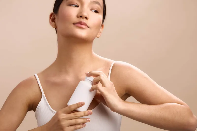 asian woman applies skincare product in a clean studio setting, wearing a white tank top. she smiles softly, presenting a calm, confident beauty routine moment.