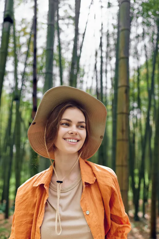 Woman smiling in bamboo forest wearing straw hat and orange jacket, outdoor portrait in nature. Young traveler enjoying a peaceful walk among tall green stalks and sunlight.