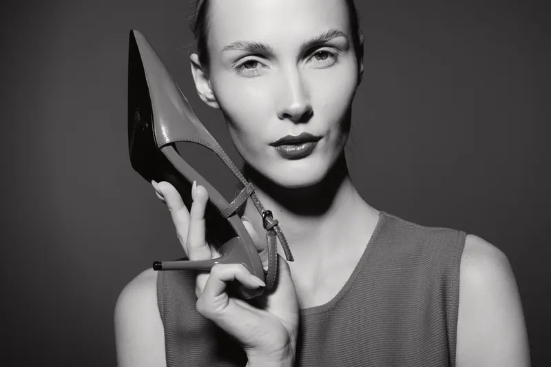Portrait of a light skinned Caucasian woman holding a high heel shoe close to her face in black and white.