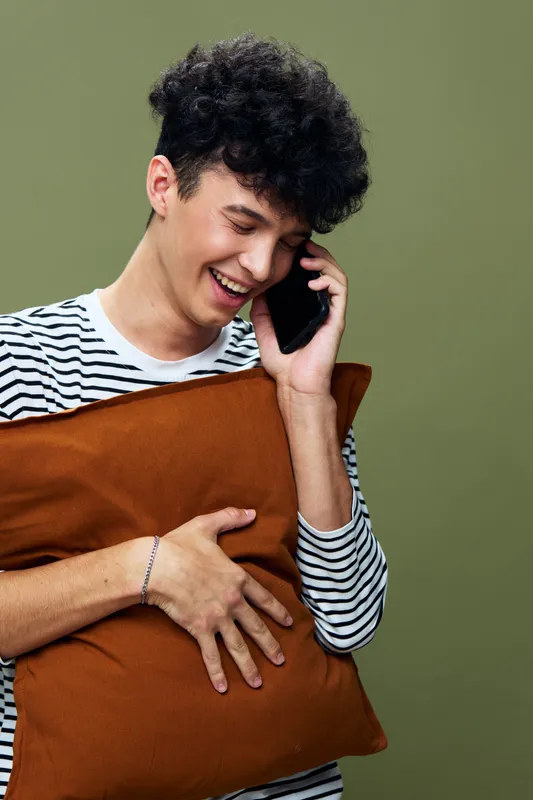 young man talking on phone smiling happily holding brown pillow casual striped shirt olive background vibrant modern lifestyle scene