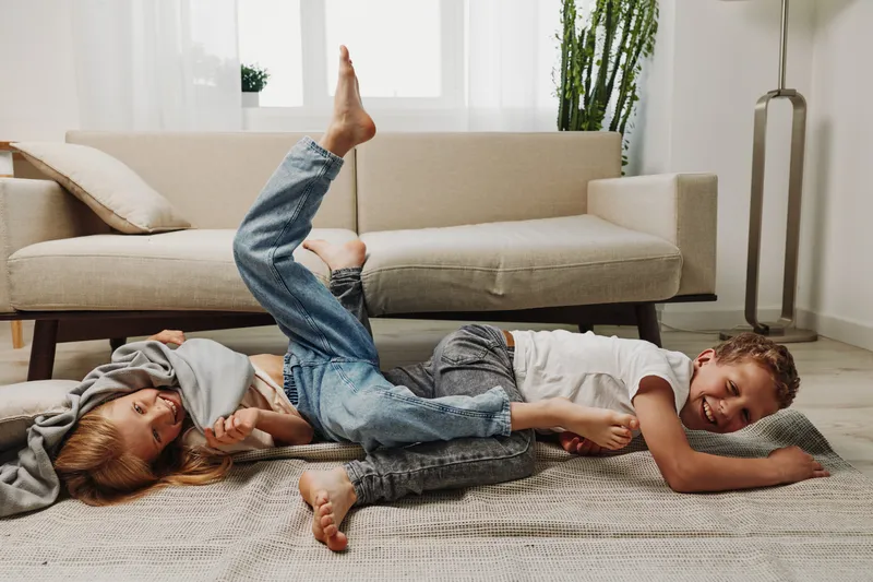 Two children in casual clothes play and laugh on the carpet in a bright living room with a sofa and plants in background.