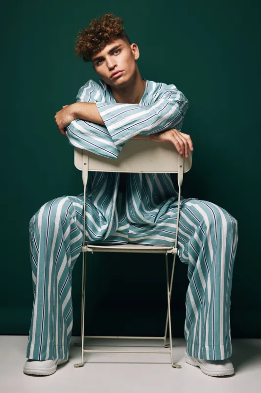 Young man with curly hair wearing striped pajamas sitting on chair backwards against dark green background in studio. Casual pose, relaxed mood and fashion style with comfortable outfit.
