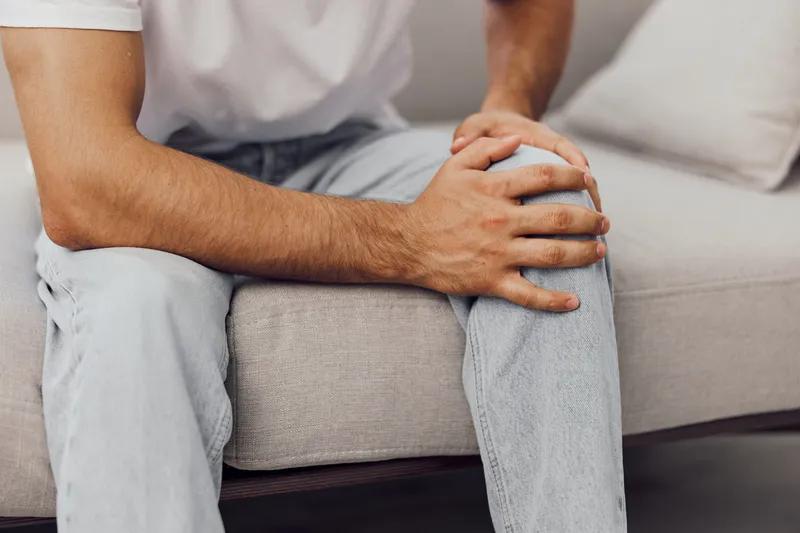 Man sitting on a couch, holding his knee in discomfort, showcasing modern home interior and neutral colors. Ideal for health and wellness themes.