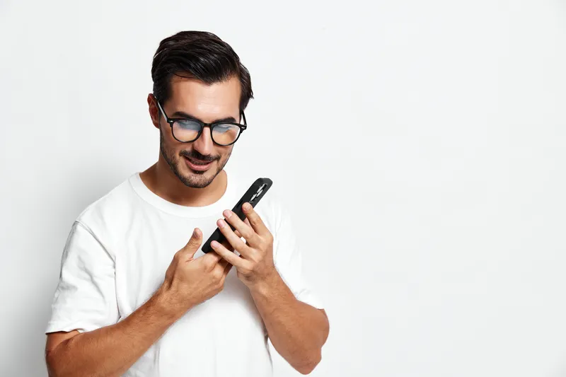 Smiling man wearing glasses holds mobile phone, looking at the camera with a cheerful expression on his face, isolated on plain white background. Happy vibe.