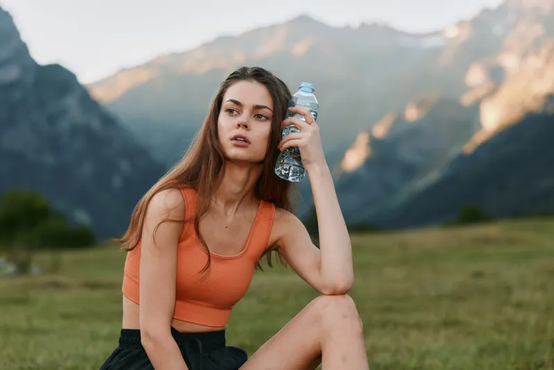 A young woman sits outdoors in a scenic valley wearing an orange top, holding a water bottle. She gazes into the distance with calm focus, enjoying nature and serenity.