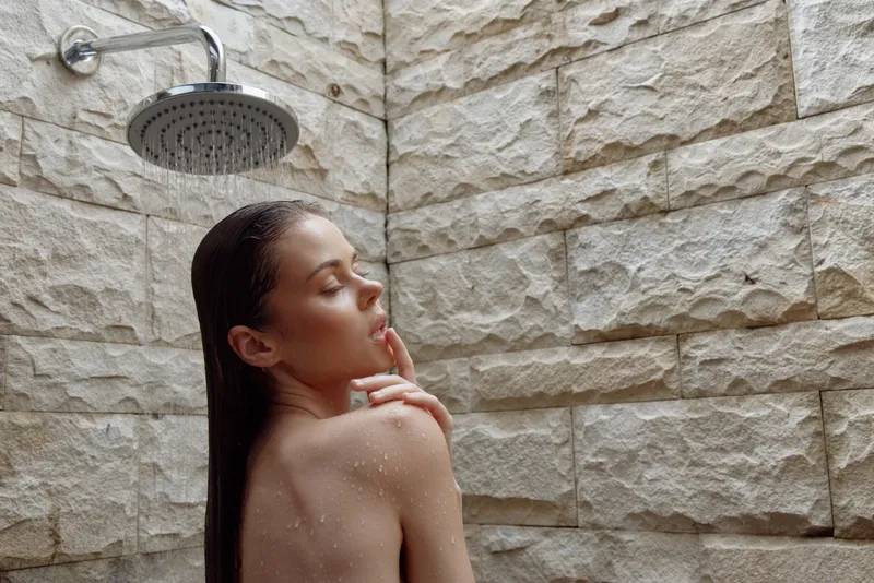 woman showering in bathroom, wet hair, relaxing under rain showerhead, natural stone wall background, calm and peaceful atmosphere