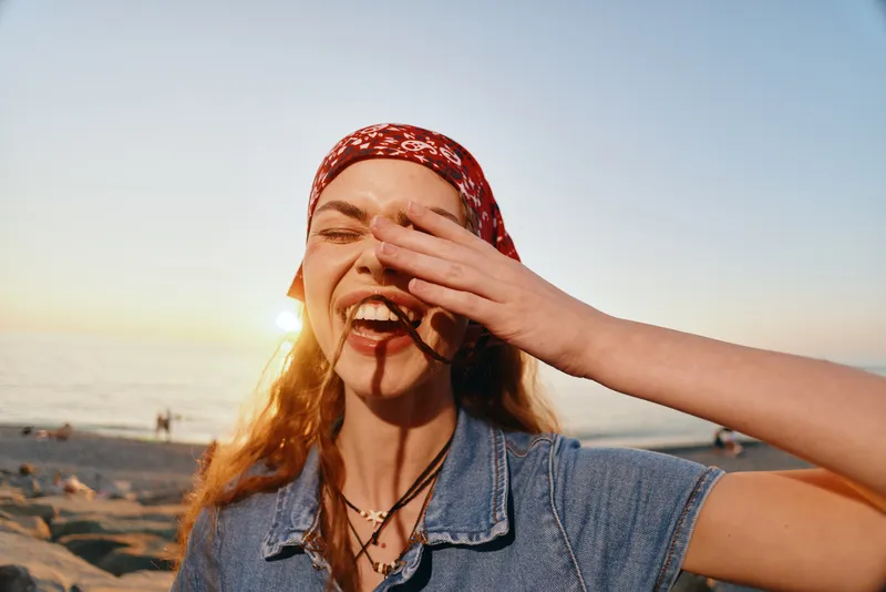 lifestyle woman laughing in modern boho-western denim street style wearing a red bandana with warm film color tones at the beach during sunset
