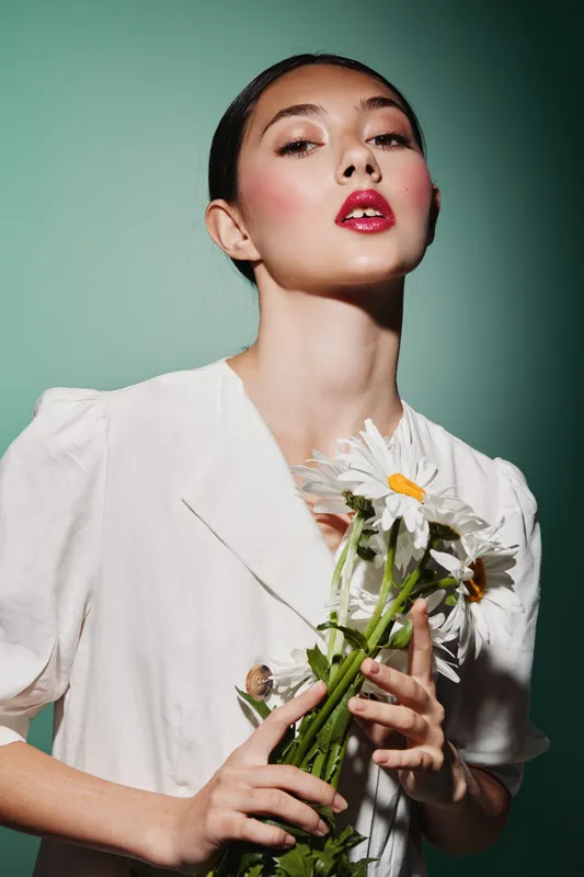 Portrait of a fashionable woman with glossy red lips holding daisies against a soft green background. Concept of beauty, fashion, floral, and creative styling in studio lighting.
