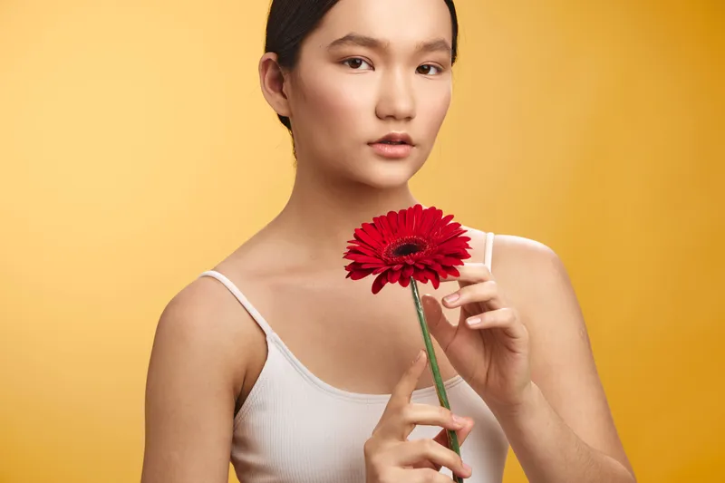 Asian woman holds a red flower in a simple studio portrait, softly lit against a warm yellow background, conveying gentle elegance and calm, natural beauty.