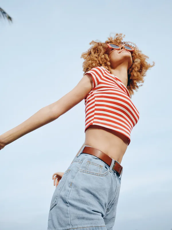Dynamic portrait of a joyful person with curly hair, wearing a red striped crop top and denim, arms extended, under bright sky, radiating freedom and energy.