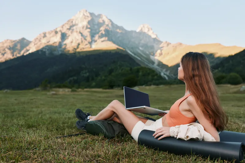 A relaxed woman sits in a grassy meadow with a laptop propped on her legs, gazing toward distant mountains, creating a serene and productive outdoor workspace.
