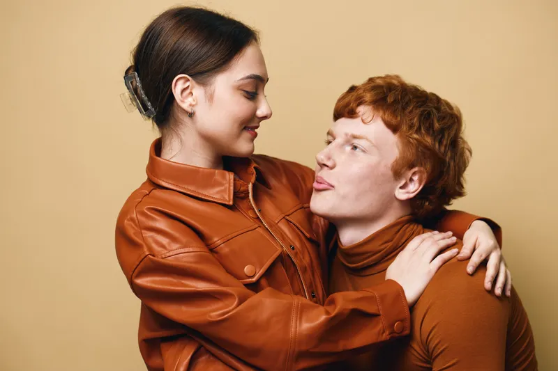 Couple, love, romance and affection with young woman and man embracing indoors. People looking at each other with smile, casual style and warm clothing on beige background.