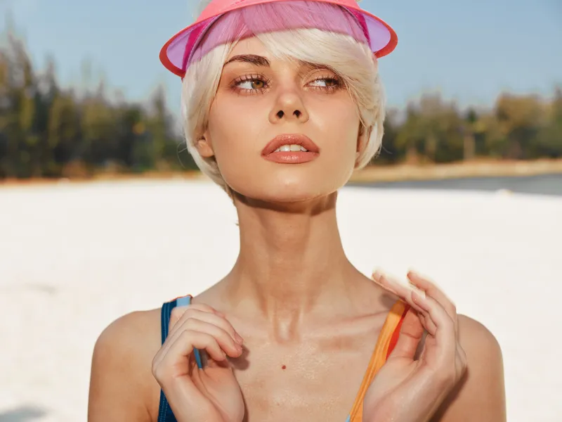 Close-up portrait of a stylish woman on a sunny beach wearing a pink visor, capturing sunlit skin, soft makeup, and a thoughtful gaze.
