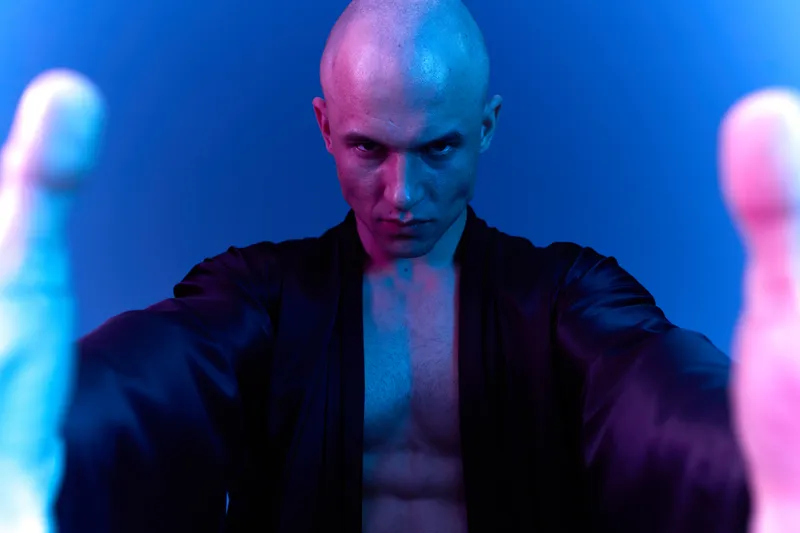 Intense man with a focused expression, dressed in a dark robe, showcasing strength and determination under blue and purple lighting.