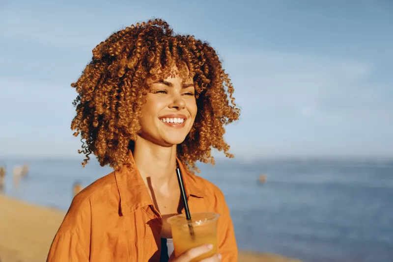 Young woman with curly hair smiles joyfully while holding a refreshing drink on a sunny beach with calm sea in the background, enjoying a relaxed summer moment.