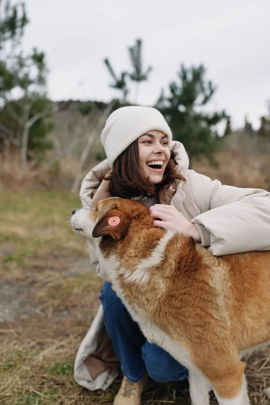 Woman in a warm coat and beanie hugs a corgi outdoors, smiling joyfully as the playful dog enjoys a moment in a chilly field, surrounded by nature and quiet countryside.