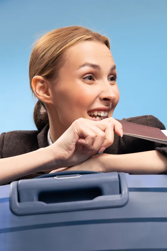 Happy woman holding passport and smiling leans on blue suitcase against light blue background. Concept of travel, vacation, tourism, and holiday preparation with positive emotions and excitement.