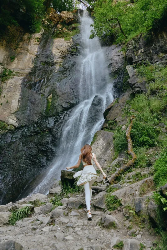 woman hiking waterfall, young woman exploring rocky nature trail near cascading water surrounded by green foliage, adventure and outdoor activity in forest