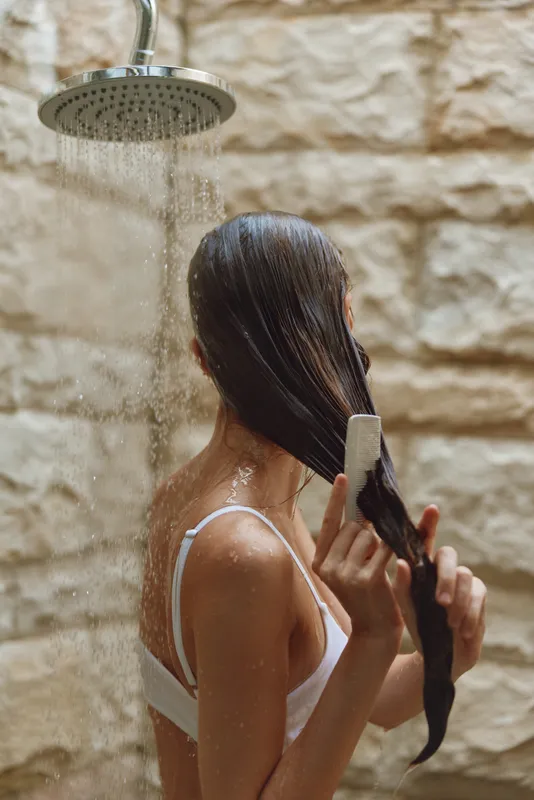 woman washing hair in outdoor shower with stone wall background, natural skin tone, long wet hair, casual white top, serene mood in warm lighting