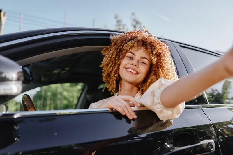 Smiling woman with curly hair leaning out of black car window enjoying sunny day outdoors with casual white shirt, bright natural light, and green trees in background.