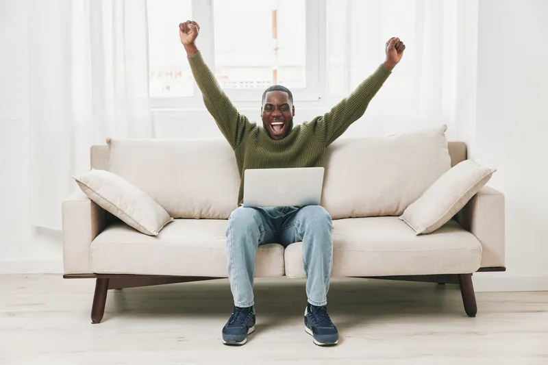 Happy African American man celebrating success while working on a laptop at home, dressed casually in a green sweater and light jeans