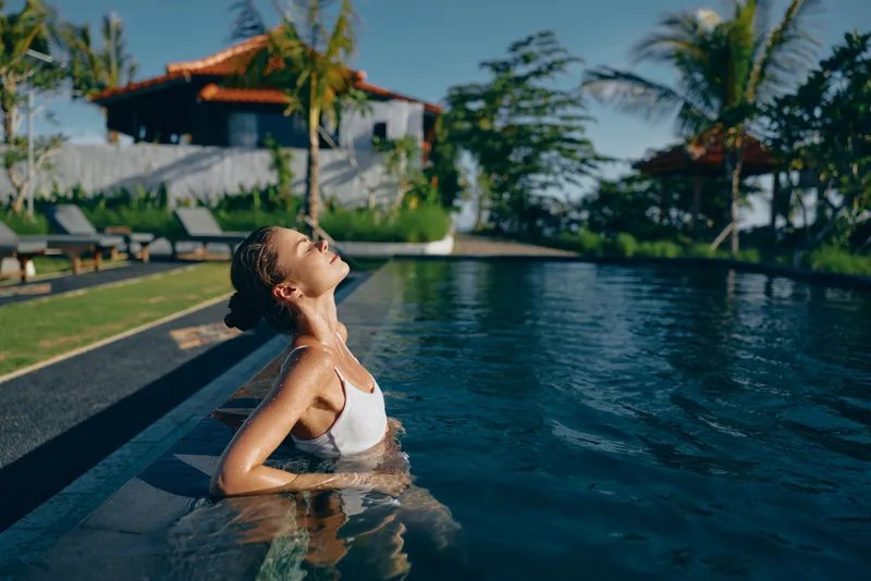 Young woman with braided hair relaxes in a swimming pool near a tropical villa, enjoying the warm sunlight and peaceful atmosphere on a summer day.