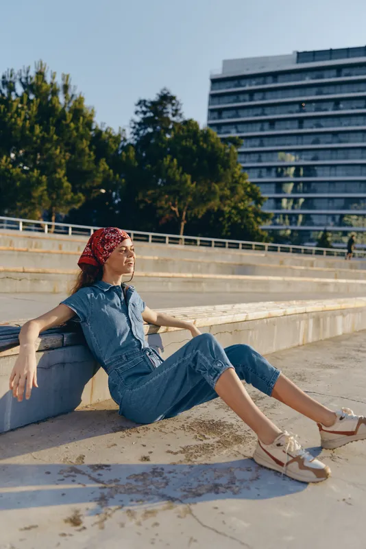 Lifestyle woman in modern boho-western denim street style sits relaxed outdoors, enjoying sunlight with casual sneakers and a red bandana, film color.