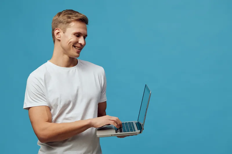 Young smiling man wearing casual white T-shirt holding a laptop computer, isolated on plain blue background, studio portrait. Office work, technology, freelance, modern lifestyle, cheerful, communicat