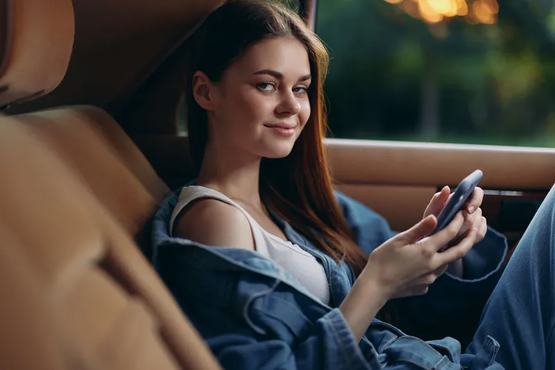 Young woman in casual denim attire relaxing in a car, smiling while using a smartphone, capturing a warm and carefree moment in nature.