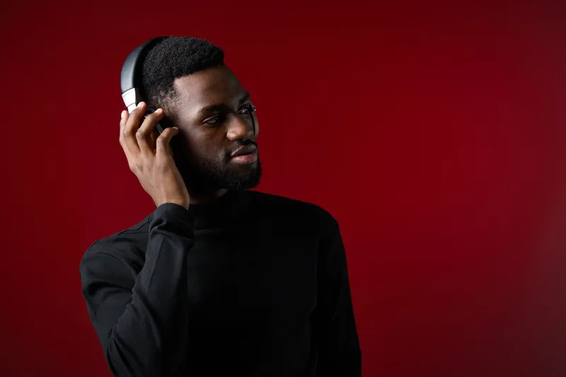 Stylish young Black man wearing headphones, enjoying music with a thoughtful expression against a vibrant red background