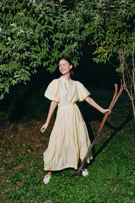 cheerful woman in a yellow dress stands in a lush garden at night holding a rake and smiling creating a warm dreamy outdoor scene with soft lighting