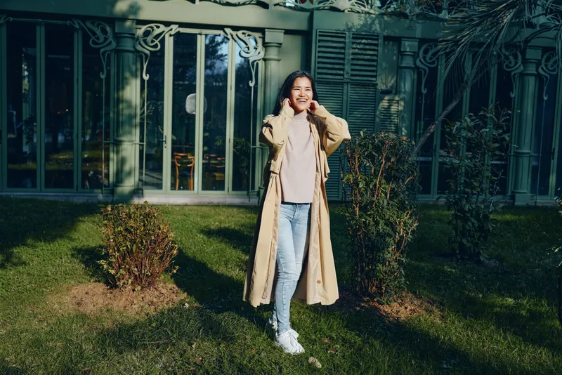 Woman wearing a long beige coat outdoors in a sunny garden, standing on grass near a glass conservatory, casual fashion and lifestyle moment in a peaceful suburban setting