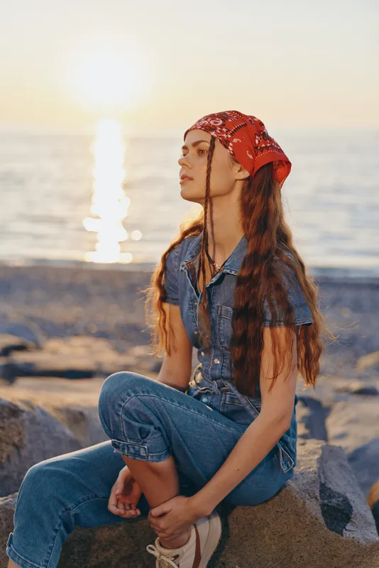 Lifestyle woman in modern boho-western denim street style sitting by the sea during sunset, wearing a red bandana, film color tones creating warm atmosphere.