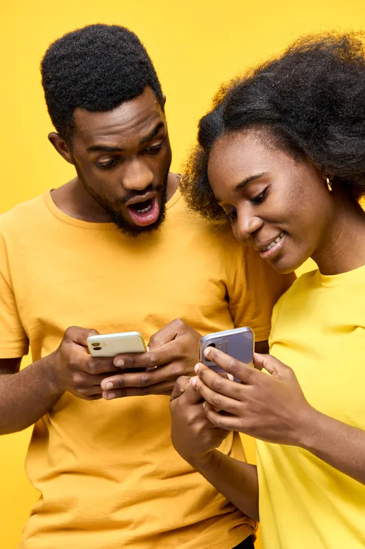 Excited young couple sharing good news on smartphones against a bright yellow background, showcasing modern communication and happiness