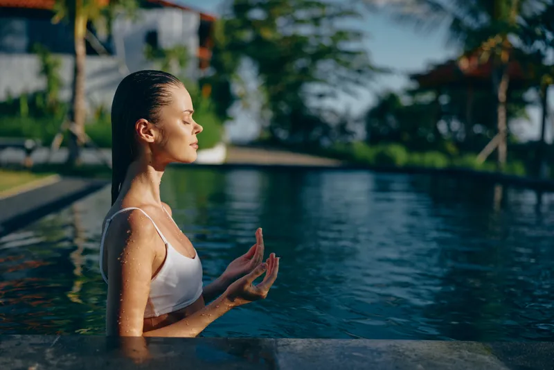 Young woman practices meditation in swimming pool at sunrise, calm and relaxed with closed eyes, enjoying peaceful lifestyle and wellness outdoors in tropical environment.