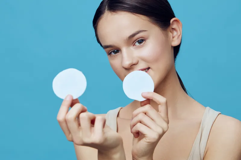 Young woman enjoying skincare routine, holding cotton pads against a blue background, promoting healthy skin and natural beauty