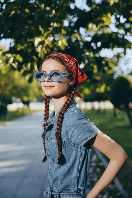 smiling woman with braids wearing blue sunglasses and red bandana outdoors in sunny park with green foliage and casual denim outfit