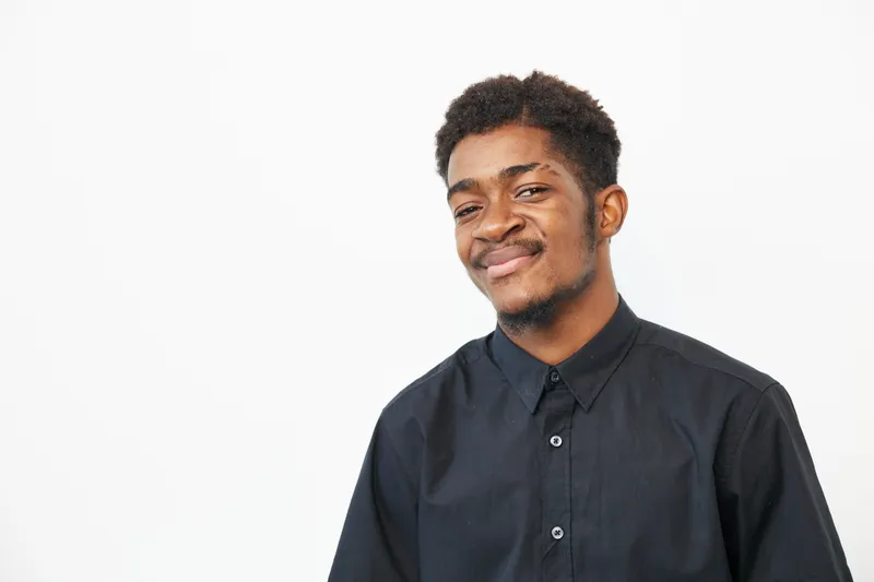 A black man with a friendly smile wearing a black shirt, exuding confidence and positivity against a clean, bright background. Perfect for lifestyle themes.