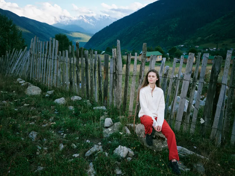 Woman outdoor fence mountains valley landscape, sits beside a weathered wooden fence in a lush valley with distant mountains, wearing red pants and a light top, capturing a calm outdoor moment in