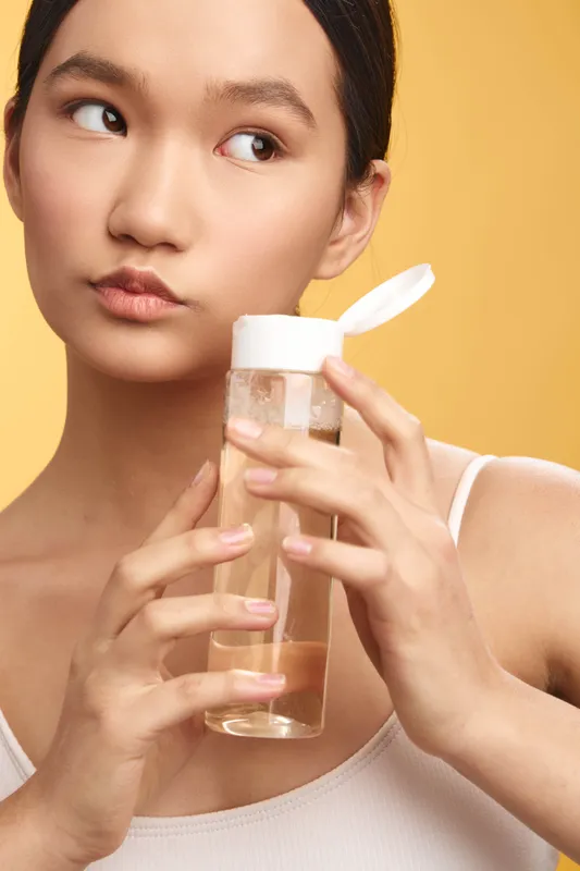 asian woman holds a clear skincare bottle near her face in a bright studio, expressing calm and focus as she demonstrates a simple beauty routine and mindful self care.