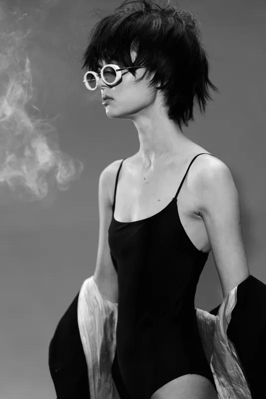 Black and white portrait of a stylish woman in sunglasses wearing a black dress and light jacket exhaling smoke in a minimalistic studio setting