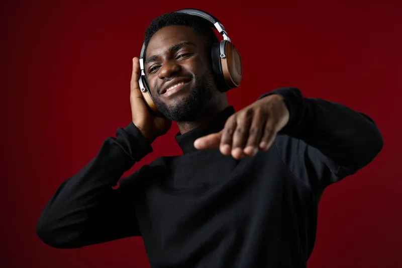 A joyful young Black man enjoys music while wearing stylish headphones against a vibrant red background, radiating happiness and a passion for sound