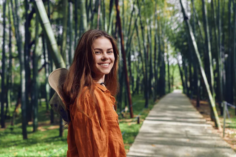 Woman smiling in a bamboo forest on a paved path, walking with a backpack and hat. Portrait of a happy traveler enjoying nature, green canopy and peaceful outdoor walkway.