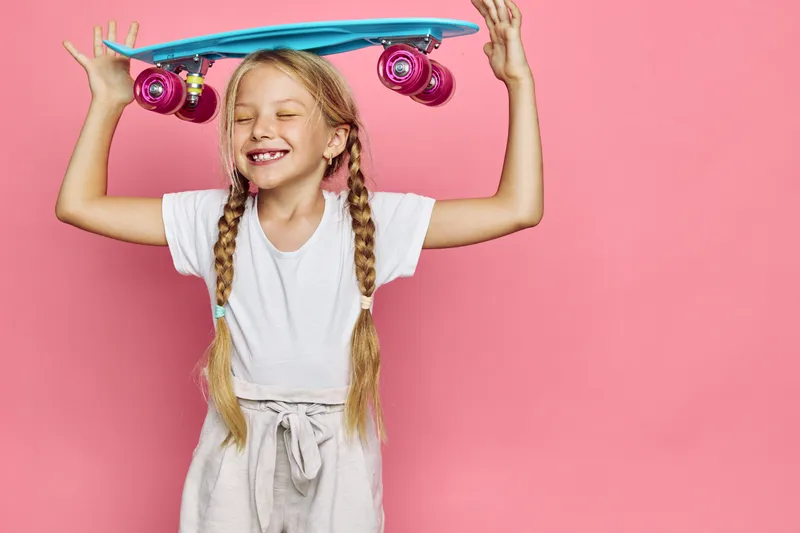 Happy child girl with braids holding blue skateboard above her head smiling with eyes closed on pink background. Concept of childhood, joy, and outdoor activities.