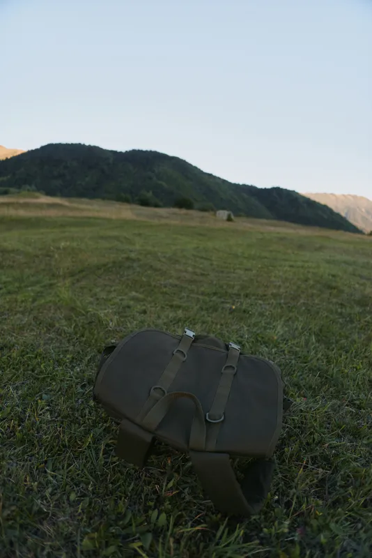 A single rustic satchel rests on a green grass field in a sunlit meadow, suggesting an outdoor escape and quiet travel mood during a calm day.
