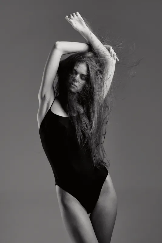 Black and white artistic portrait of a woman with long hair wearing a black bodysuit, posing elegantly with arms raised and eyes closed