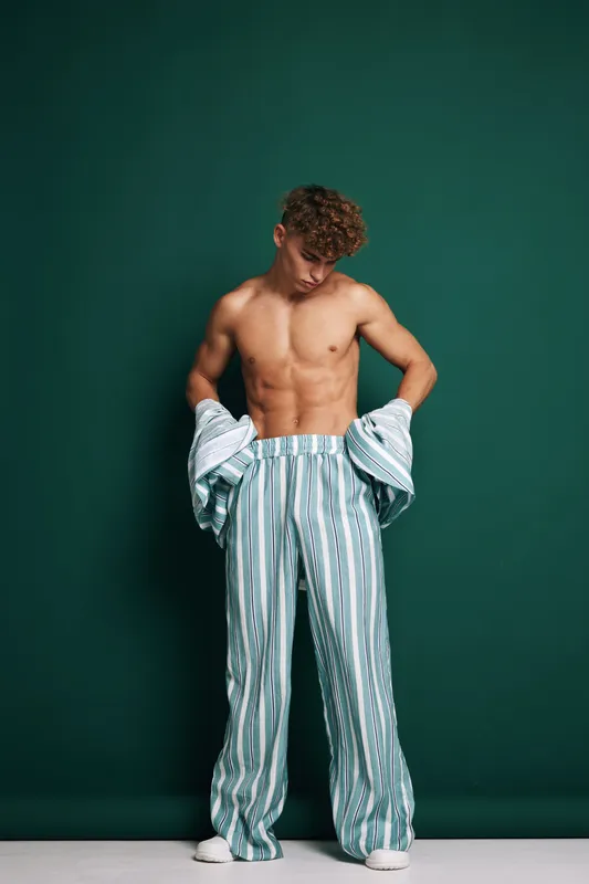 Young man with curly hair and bare torso wearing striped loose pants and holding matching shirt in studio with green background. Casual fashion and relaxed pose for style concept.