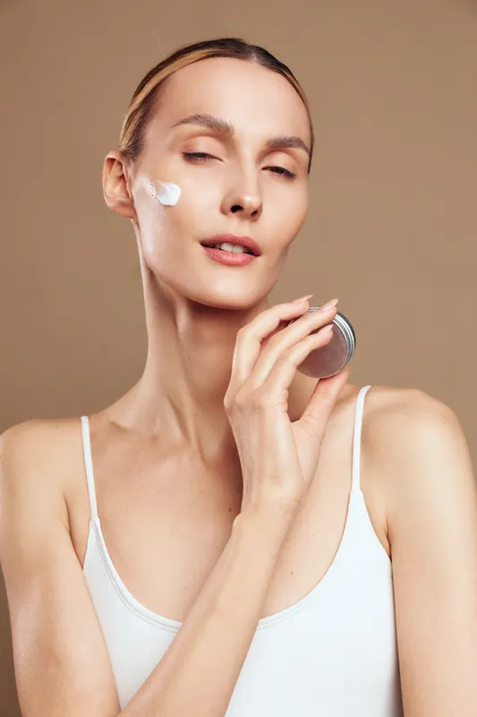 Smiling woman with smooth skin applying white cream on her cheek. Close up on a neutral background. Skincare routine, facial care, beauty ritual, self care moment, soft studio lighting.